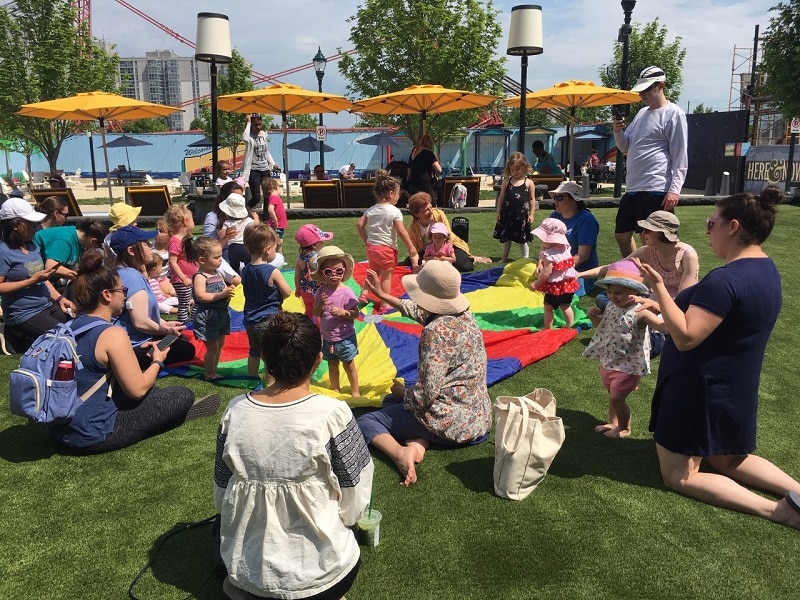 Group of families with young children at program outside on grass with umbrellas and playtime parachute.