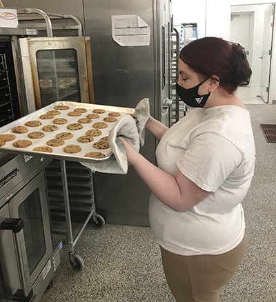 Photo of Jolie, a Sunflower Bakery employee putting tray of cookies into oven in professional kitchen.
