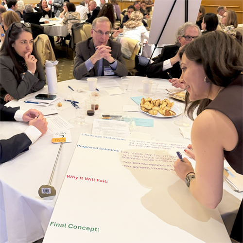 HARTMA~1 Group of three community leaders during Hartman Convening March 2025 at table with Ari Brickman writing barinstorming notes on poaster board with topics for discussion Challenge Statement, Proposed solution final concept