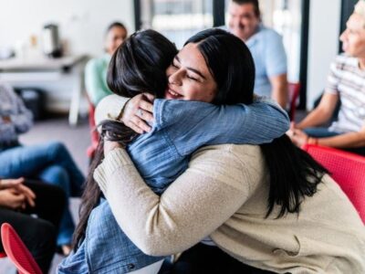 251ef83aa503f5af270b4782 Two young women hugging with people sitting around them in a circle.