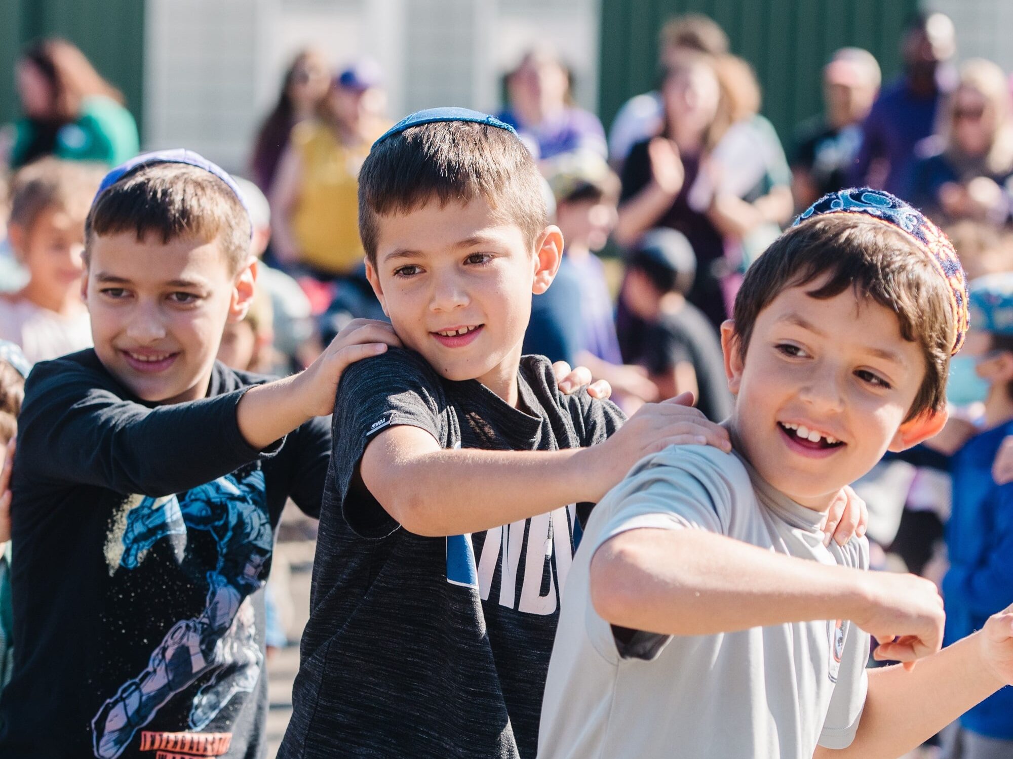 Three young boys at Gesher Jewish Day School with arms on each other's shoulders in a dance line.
