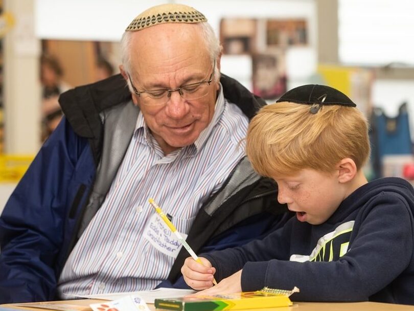 A grandfather and his grandson, both wearing kippot, sit at a desk in a classroom