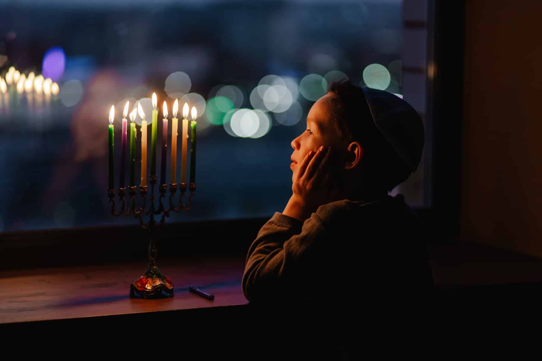 A little boy looking at a menorah sitting by a window
