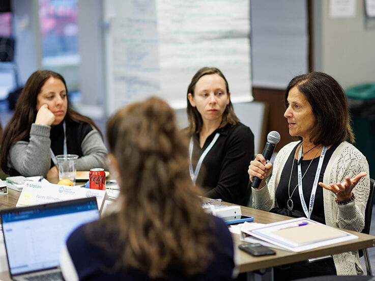 Group of people sitting at table, while one woman with brown hair is speaking into a microphone