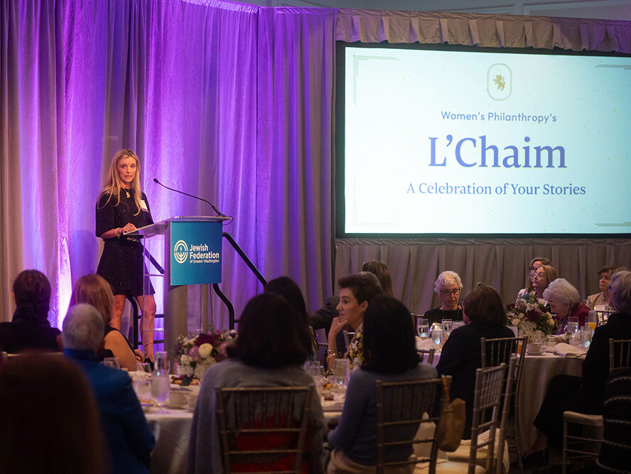 Woman speaking at a podium with the Jewish Federation of Greater Washington logo, addressing an audience at round tables during the Women's Philanthropy L'Chaim event, with purple lighting and a large screen displaying event details.