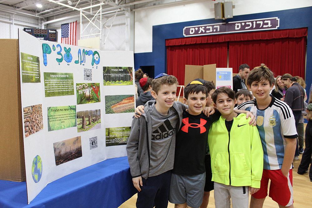 Four boys stand together at school in front of a home made poster displaying images of Israel and Hebrew lettering.