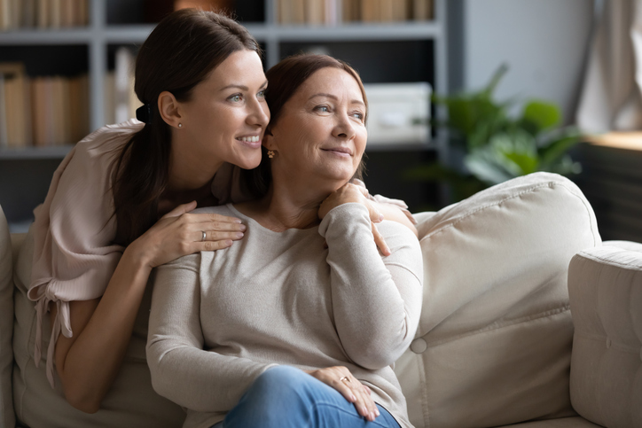 Smiling middle-aged mom and adult daughter in a cozy living room, one seated on a couch and the other standing behind with hands on the seated person's shoulders, with a bookshelf full of books and green plants in the background.