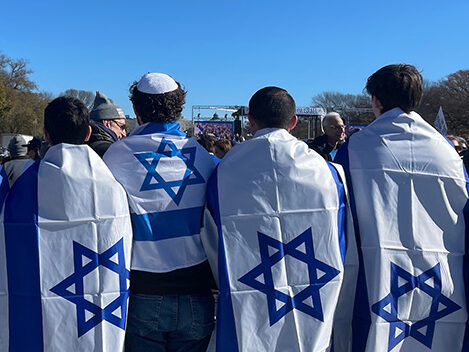 Group of people stand together, facing away, with the Israeli flag draped on them